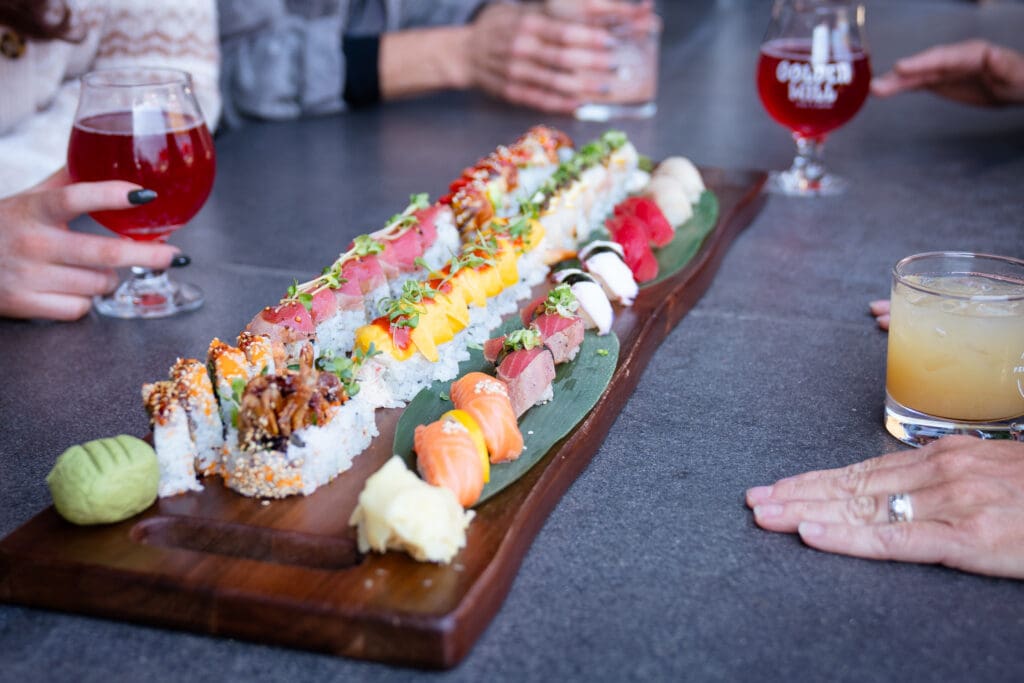Long wooden board filled with sushi rolls and nigiri, surrounded by diners and drinks at The Golden Mill.